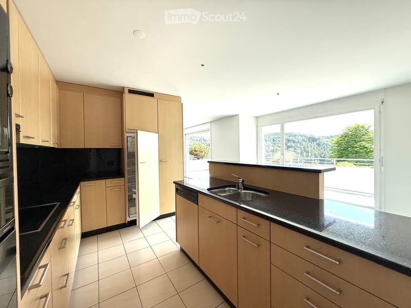 Modern kitchen with light wooden cabinets, black countertops, stainless steel sink, and appliances. Large windows offer a scenic view of greenery and a pool area.