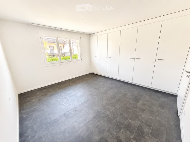 Empty room with white walls, grey tiled floor, large built-in white wardrobes, and a window with three panes showing a view of neighboring houses and greenery.
