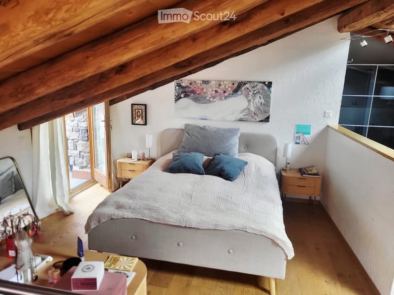 Cozy attic bedroom with wooden beams, a large bed with pillows and blankets, nightstands with lamps, a small table with books and decorative items, and artwork on the walls.
