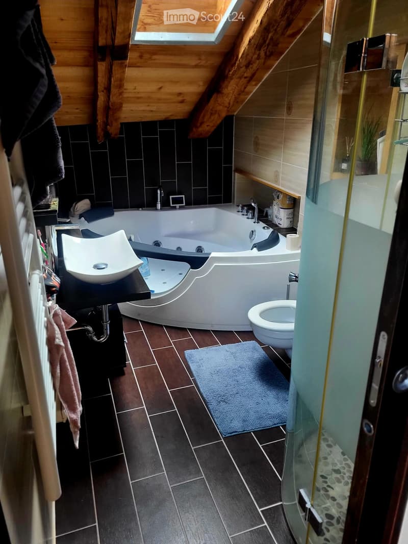 Modern bathroom with a large bathtub, sink, toilet, and a glass-enclosed shower area. The room features dark tiled floors and walls, a wooden slanted ceiling, and a small shelf with cleaning supplies.