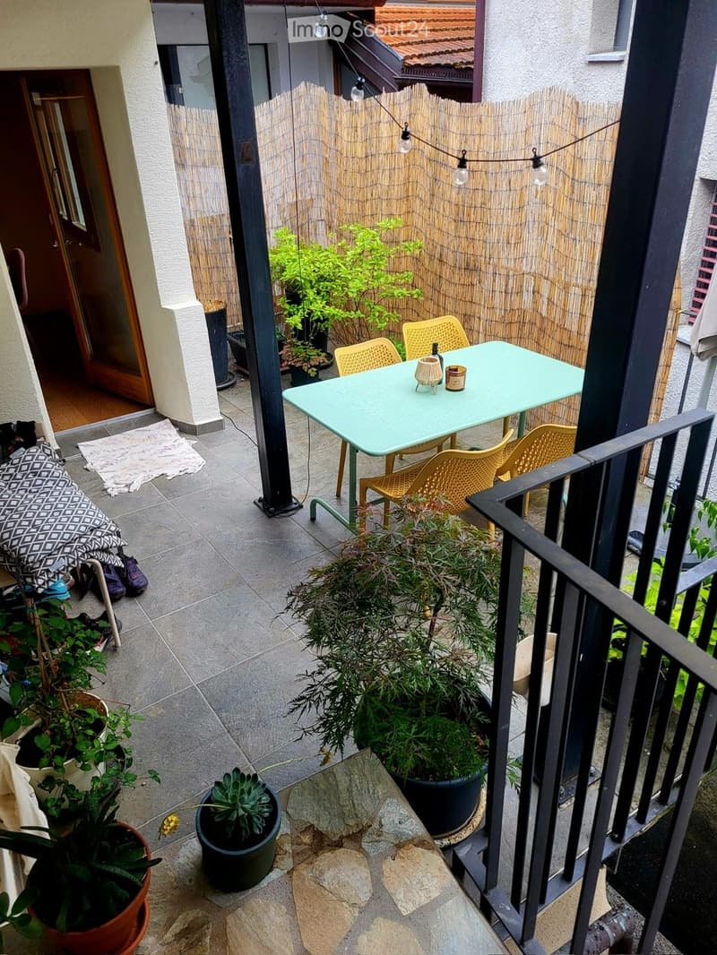 Balcony with a small table, chairs, and several potted plants. The area is enclosed by a bamboo screen and has a tiled floor.