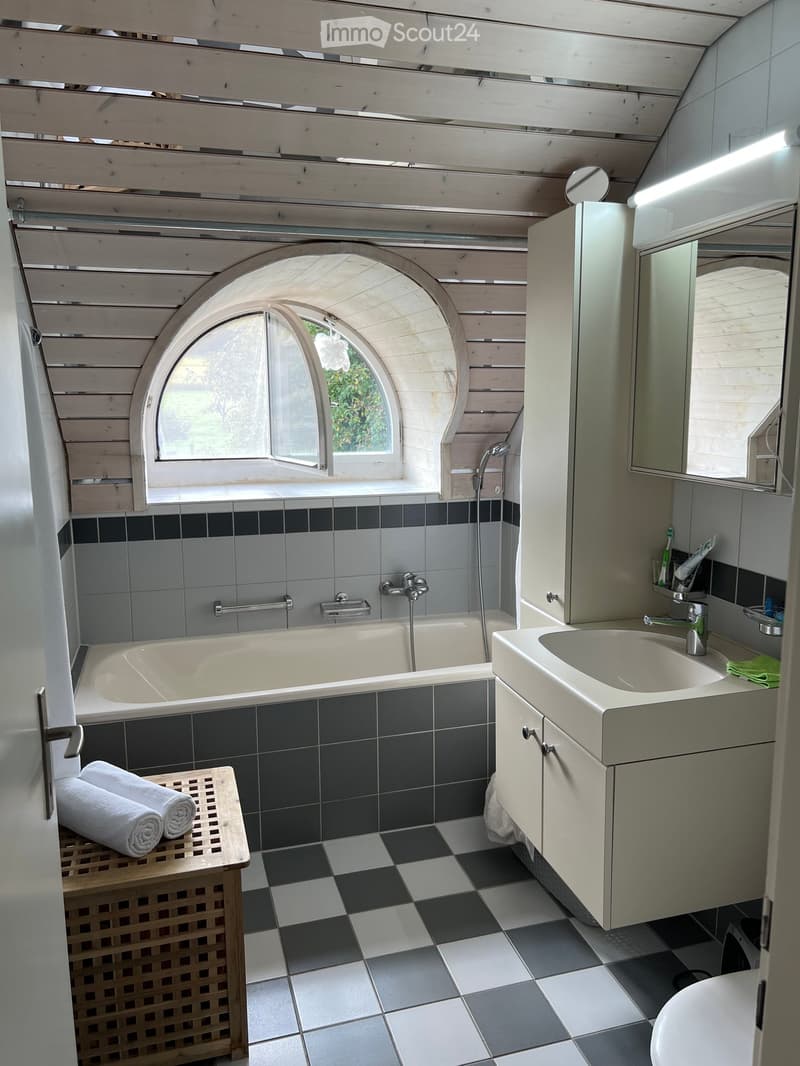 Bathroom with a bathtub, sink, mirror, and cabinet. Features a large arched window, checkered floor, and a small wooden shelf with towels.