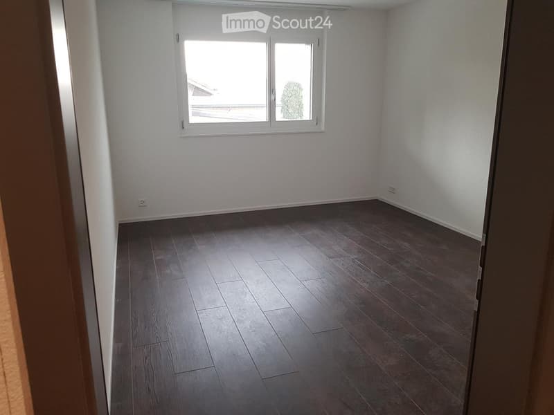 Empty room with wooden flooring and white walls, featuring a window with white frames and a view of greenery outside.