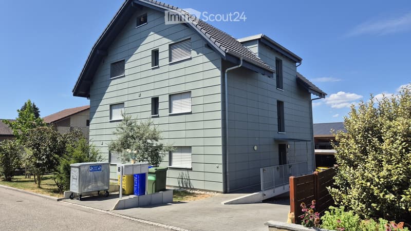Two-story house with grey siding, multiple windows, and a small garden area with a bench and plants.