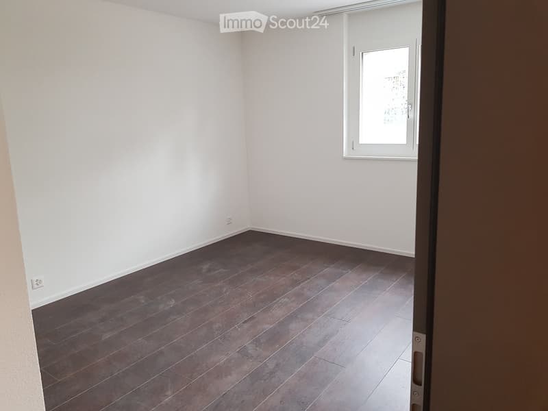Empty room with dark wooden flooring and white walls, featuring a window with blinds and an open doorway.