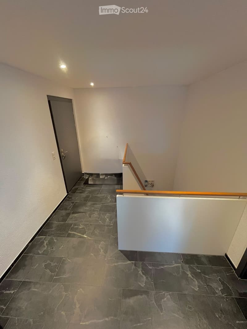 Empty hallway with dark tiled floor, white walls, and a wooden handrail leading to a door at the end.
