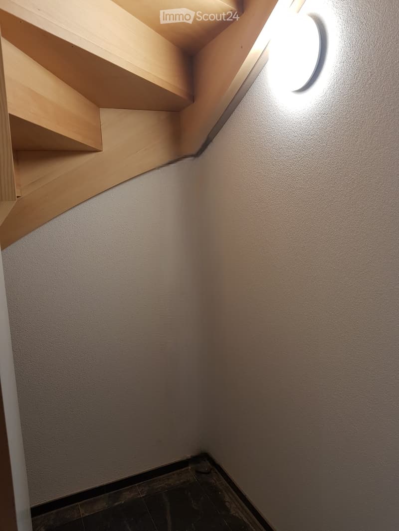 A narrow staircase with wooden steps and white walls, illuminated by a wall-mounted light fixture.