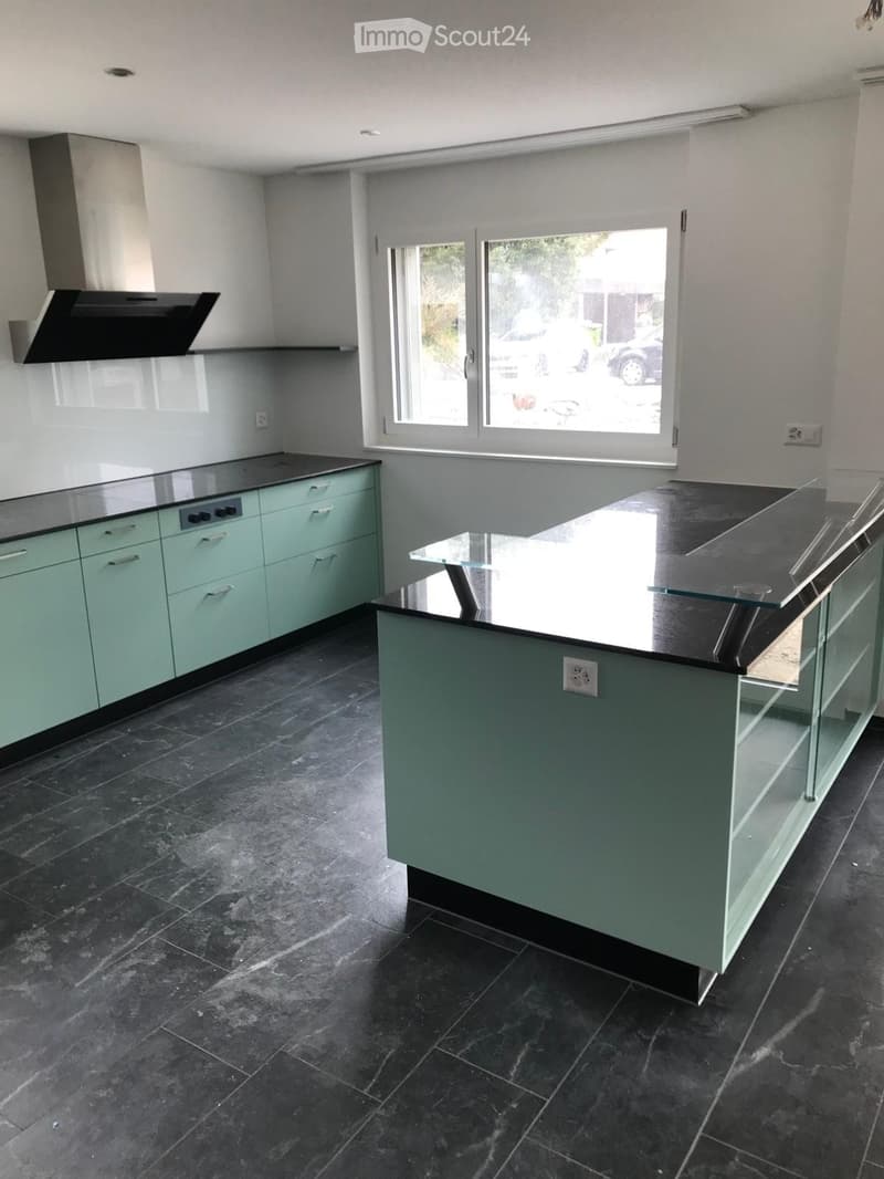Modern kitchen with light green cabinets, black countertops, and a central island with glass front. Features a range hood and large windows letting in natural light.