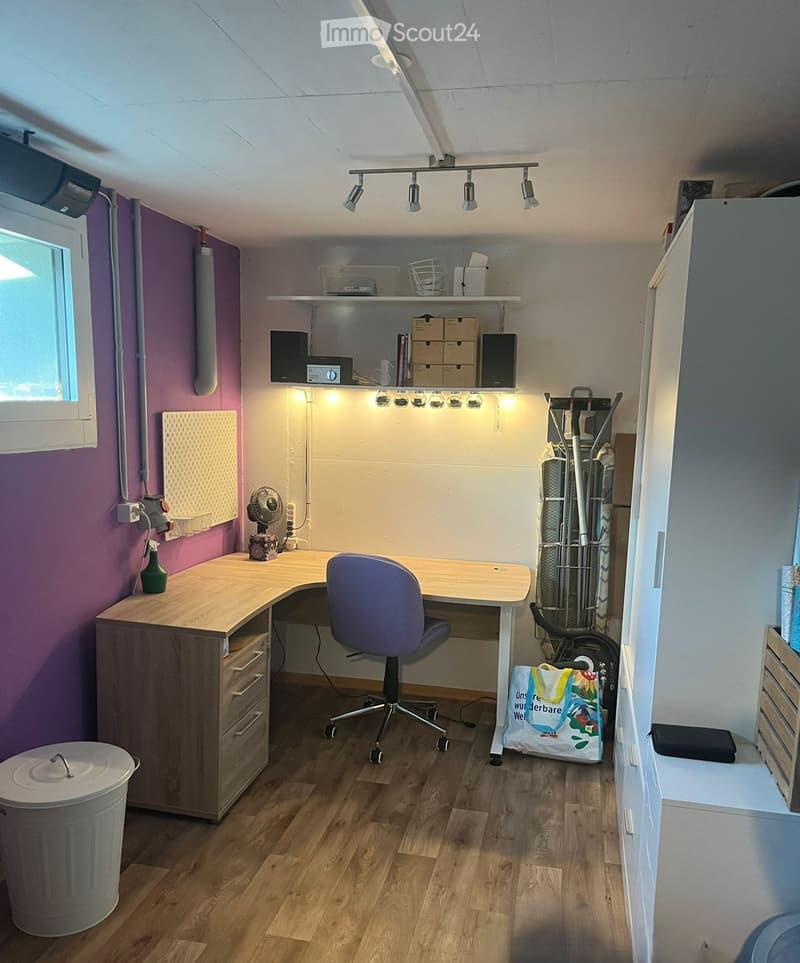 A well-lit study or office space with a wooden desk, ergonomic chair, fan, speakers, storage boxes, and a trash bin.