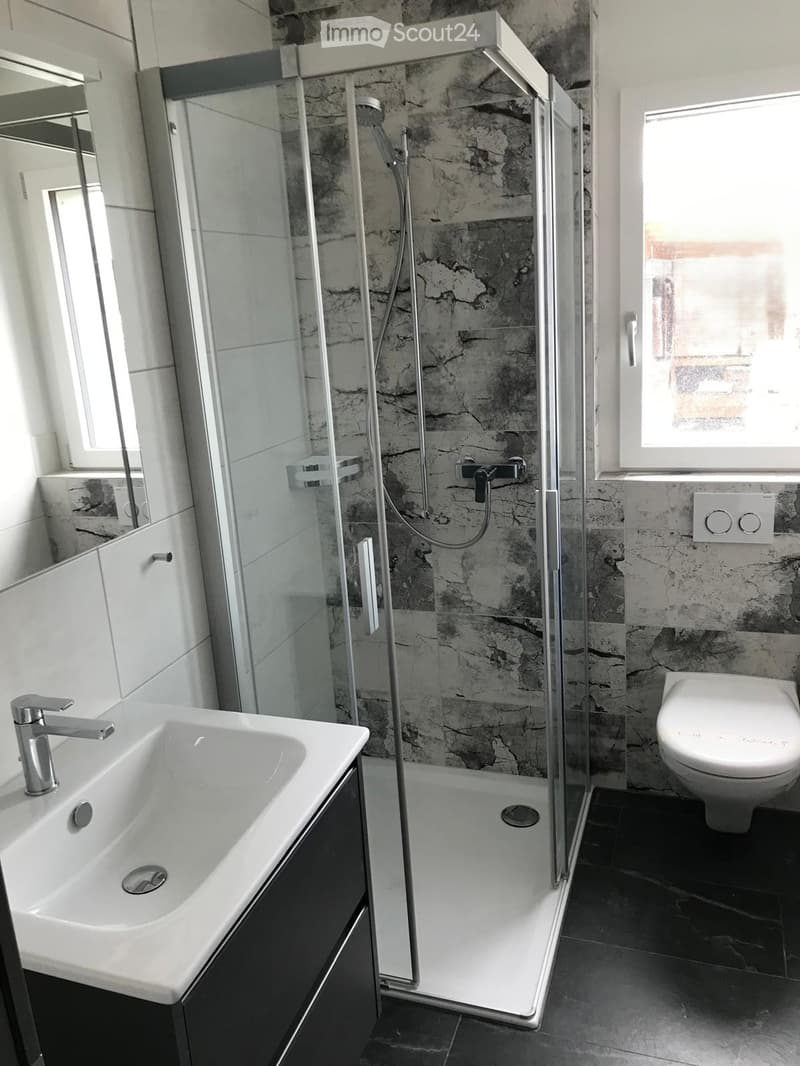 Modern bathroom with a white sink, chrome faucet, glass-enclosed shower, and toilet. The walls have a stone-like texture, and the floor is dark tiled.