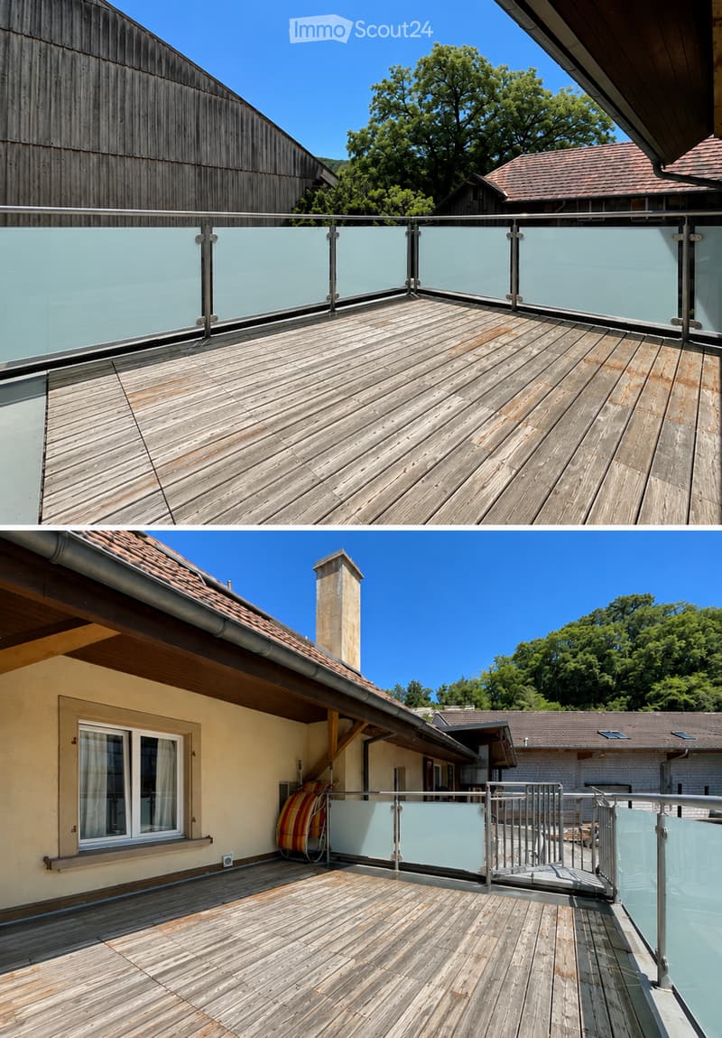 Wooden deck with glass railing, clear sky, surrounding trees, and neighboring buildings visible.