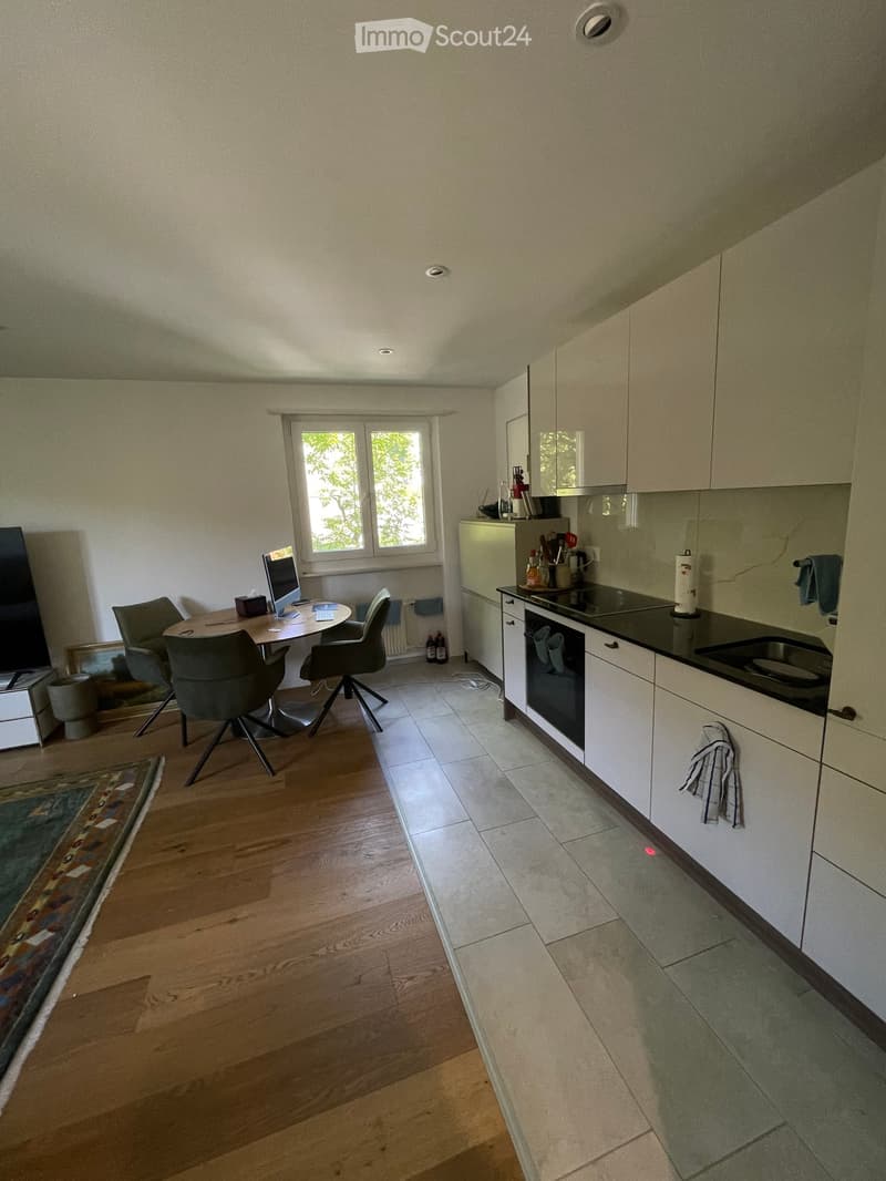 A modern kitchen with white cabinets, a black countertop, and a small dining area with a table and chairs. The floor is a combination of wooden and tiled surfaces.
