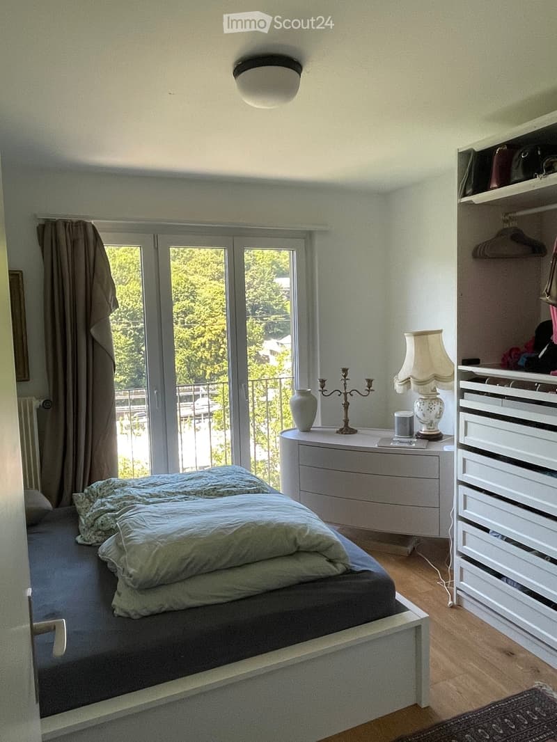 Cozy bedroom with a double bed, nightstand, lamp, vase, candle holder, and wardrobe. Large window with balcony access and natural light.