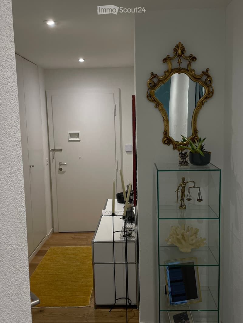 Hallway with wooden floor, glass shelf with decorative items, mirror with ornate frame, candle holder, and a door leading to another room.