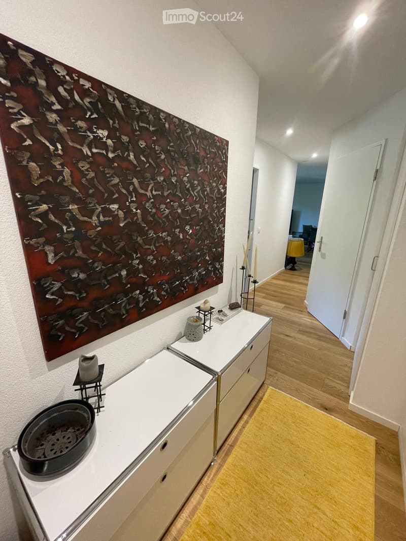 Hallway with wooden flooring, white walls, a large abstract painting, a small console table with decorative items, and a yellow rug leading to another room.
