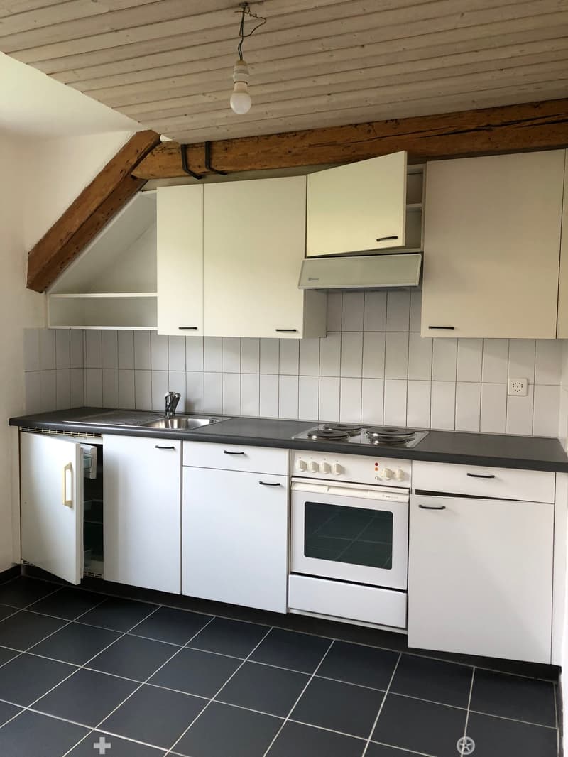 Modern kitchen with white cabinets, black countertops, and stainless steel appliances including a sink, stove, and oven. The room features exposed wooden beams on the ceiling and dark tiled flooring.