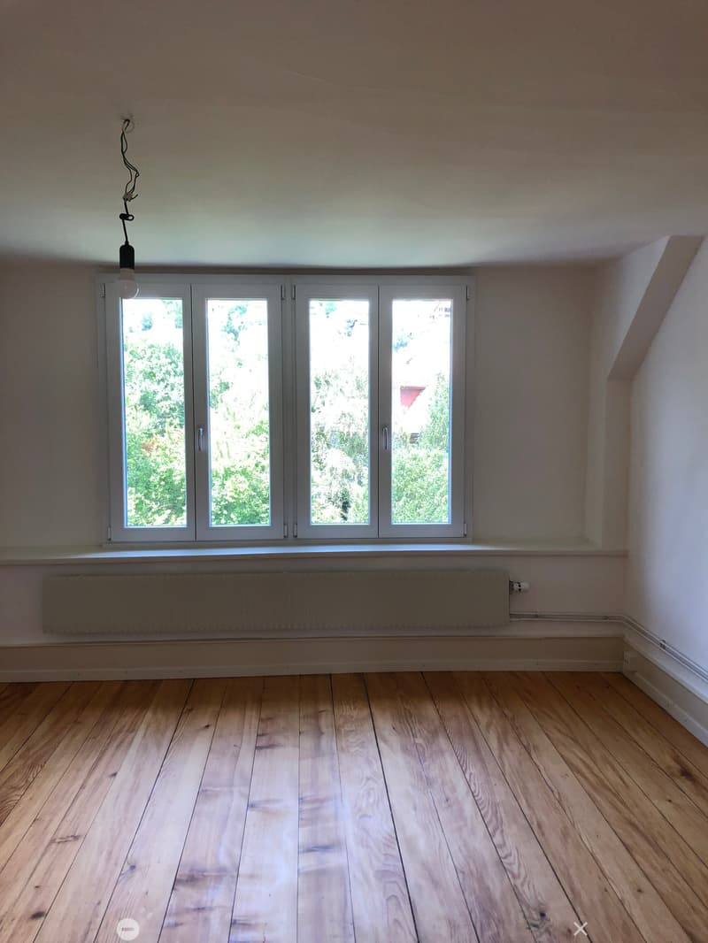 Empty room with wooden flooring, large windows, radiator, and a single hanging light fixture.