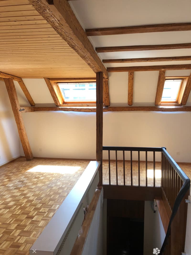 An empty hallway with wooden floors, exposed wooden beams, and skylights providing natural light. The railing is black metal, and the walls are white.