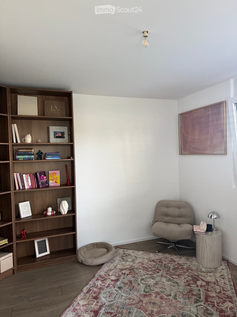 A cozy, well-furnished room with a wooden bookshelf filled with books and decorative items, a comfortable armchair, a small round table with a lamp, and a patterned rug on the floor.