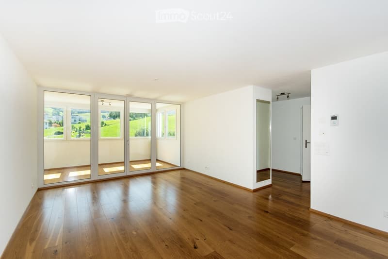 Empty room with wooden flooring, white walls, large sliding glass doors, and a view of a green landscape.