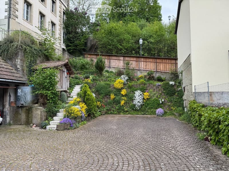A narrow, cobblestone pathway leads up a hillside garden with colorful flowers, shrubs, and trees, bordered by stone walls and wooden fences.