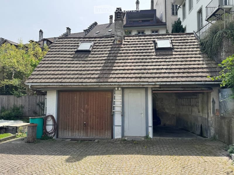 A small, two-story building with a tiled roof, featuring a wooden garage door, a white entrance door, and an open garage area. The surrounding area includes a paved courtyard and neighboring structures.