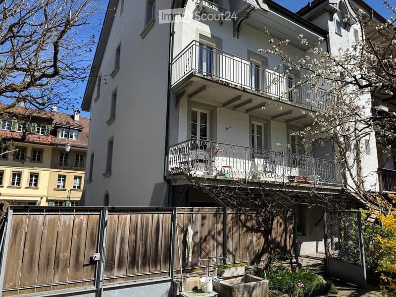 Multi-story residential building with balconies, ornate metal railings, and a fenced garden area with plants and a small sink.