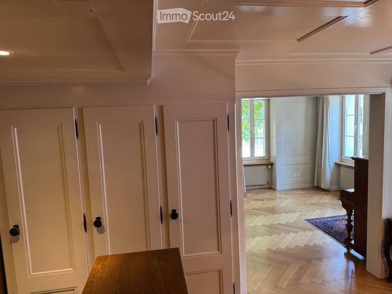 A well-lit hallway with wooden flooring, white walls, and multiple doors leading to other rooms. The hallway features elegant molding and a small wooden bench at the entrance.