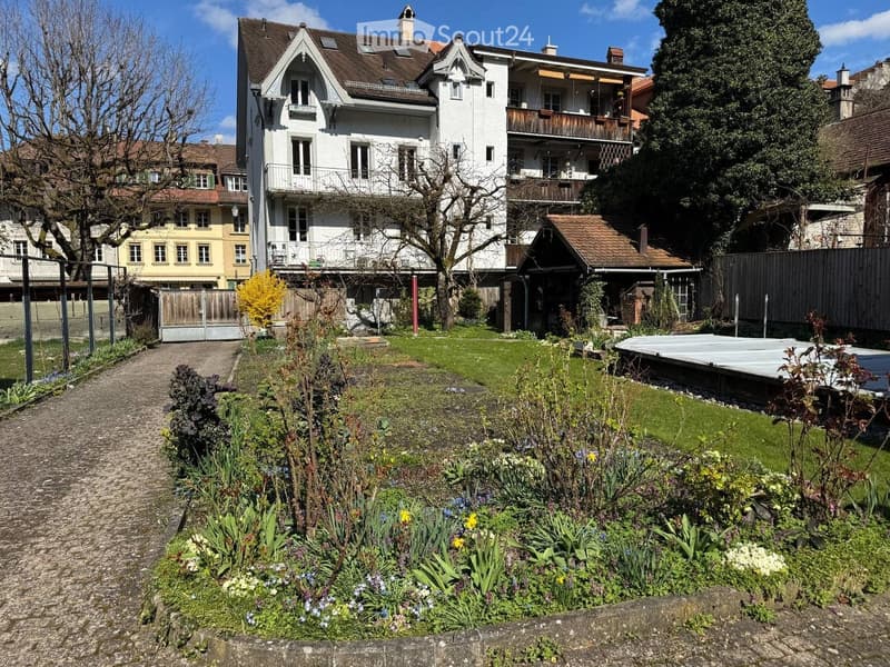 Garden with a gravel path, flower beds, and a swimming pool in the background, surrounded by trees and a large house with balconies.