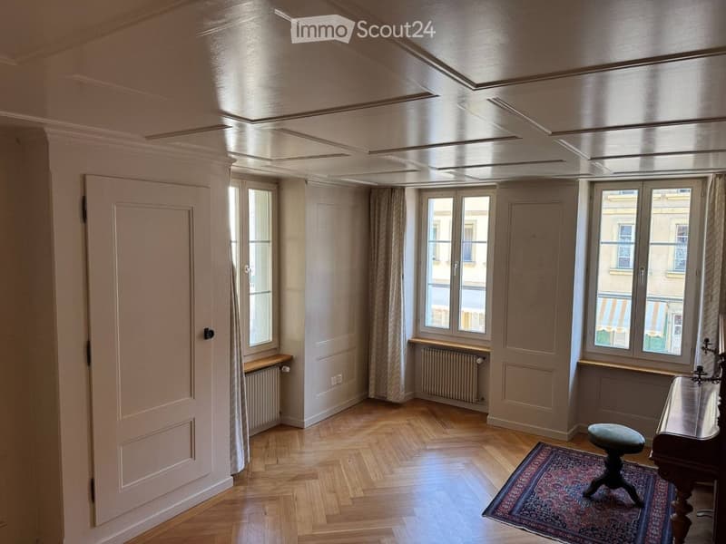 Spacious room with wooden flooring, large windows, radiators, and a decorative rug on the floor.