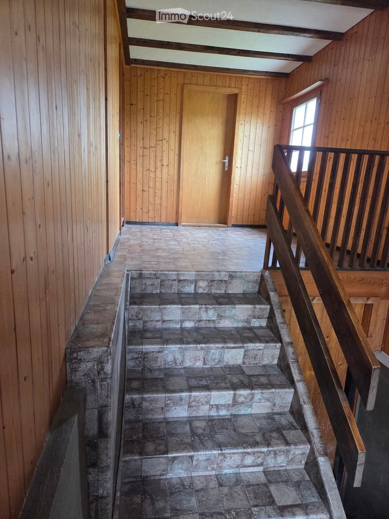 Wooden-paneled hallway with a staircase leading upwards, featuring a wooden handrail and tiled steps.