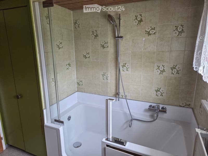 Bathtub with a glass shower enclosure, modern faucet, and handheld showerhead. Floral tile design on walls.