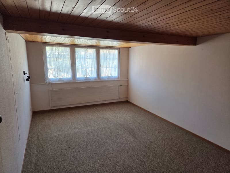 Empty room with beige walls, carpeted floor, large window with white curtains, radiator, and wooden ceiling beams.