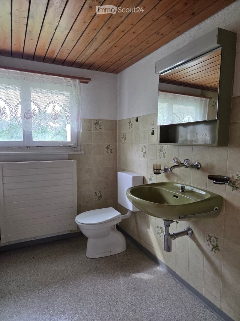 Bathroom with a toilet, sink, mirror, and radiator. The walls are tiled with floral patterns, and there is a window with a lace curtain.