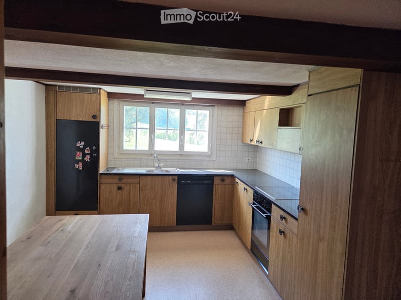 Modern kitchen with wooden cabinets, stainless steel appliances, and a large window providing natural light. Features include a refrigerator, sink, dishwasher, oven, and a wooden table.