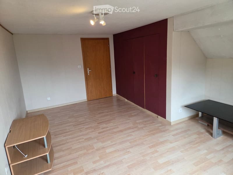 A small, unfurnished room with wooden flooring, a single wooden door, and a small wooden shelf and table.