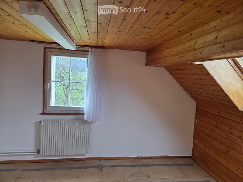 Empty attic room with wooden ceiling and walls, a window with white curtains, and a radiator below the window.