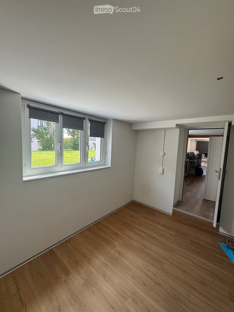 Empty room with wooden flooring, white walls, and large windows with roller shades. An open doorway leads to another room with visible furniture.