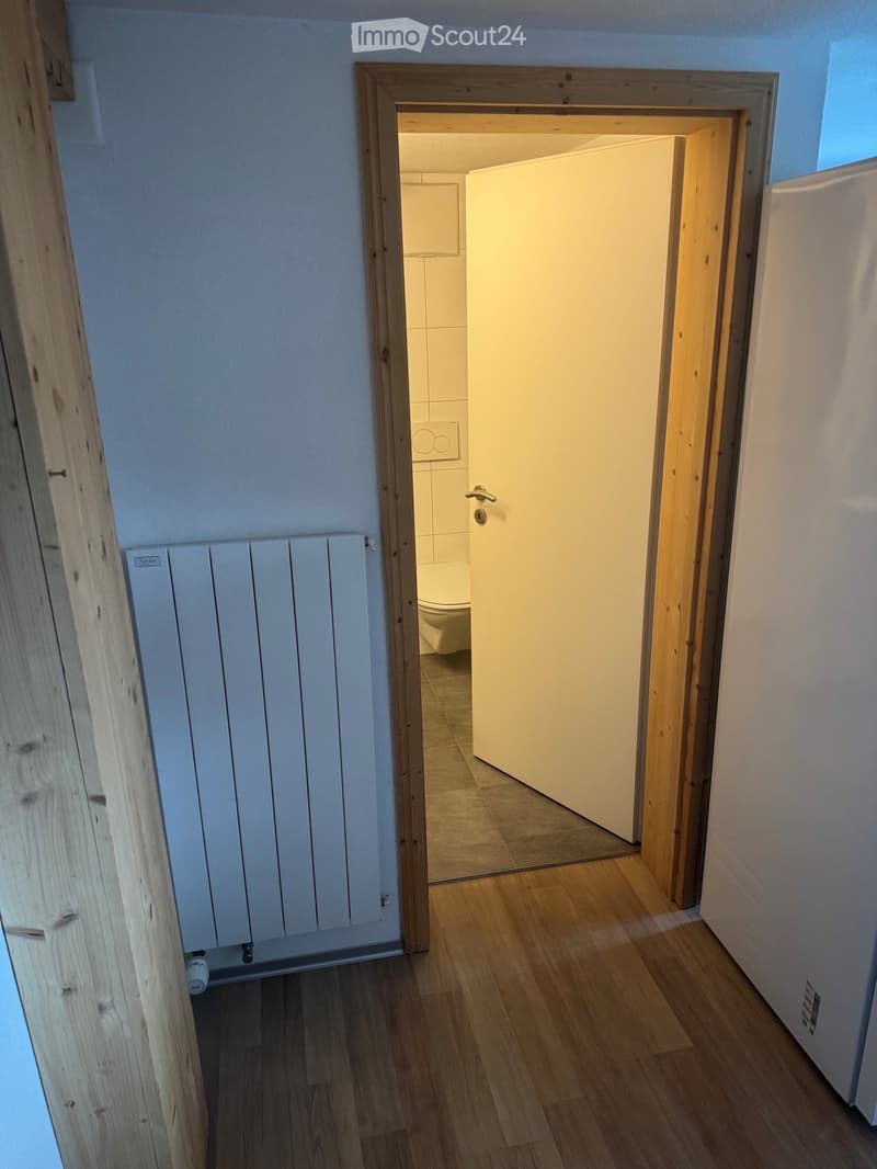 A narrow hallway with wooden flooring, white walls, and a radiator on the left side. An open door leads to a small bathroom with a toilet and tiled walls.