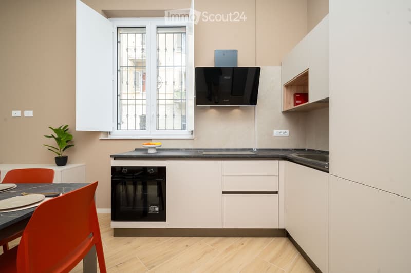 Modern kitchen with white cabinets, black countertops, built-in oven, range hood, and a small dining area with a table and chairs.