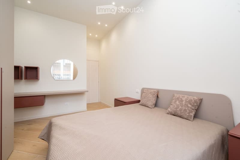 Modern bedroom with a single bed, nightstand, decorative pillows, and built-in shelves. The room features light-colored walls, wooden flooring, and a sleek, contemporary design.