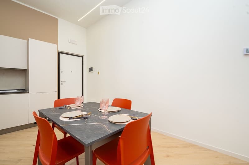 Dining area with a marble table, six orange chairs, and a set of dishes and cutlery arranged on the table. The room features white walls, light wooden flooring, and a white cabinet in the background.