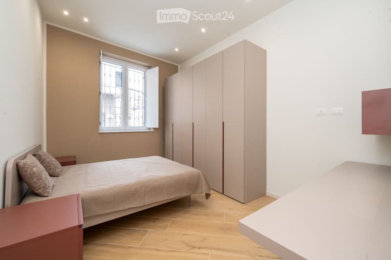 Modern bedroom with a single bed, pillows, a wardrobe, and a nightstand. The room features light wooden flooring and white walls with a large window.