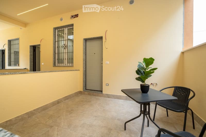 Bright, furnished room with tiled floor, yellow walls, a small black table with a potted plant, and two black wicker chairs. Features a door with a decorative metal frame and windows with metal bars.