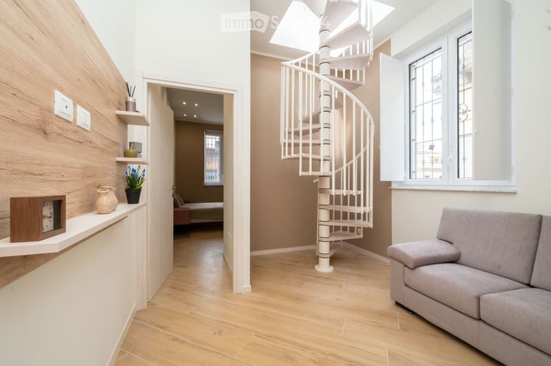 Modern living room with light wooden flooring, a grey sofa, a white spiral staircase, decorative vases, a potted plant, and shelves with decorative items.