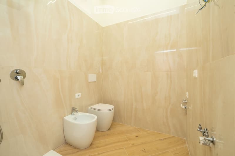 Modern bathroom with light beige tiled walls and wooden flooring, featuring a toilet, bidet, and modern faucet.