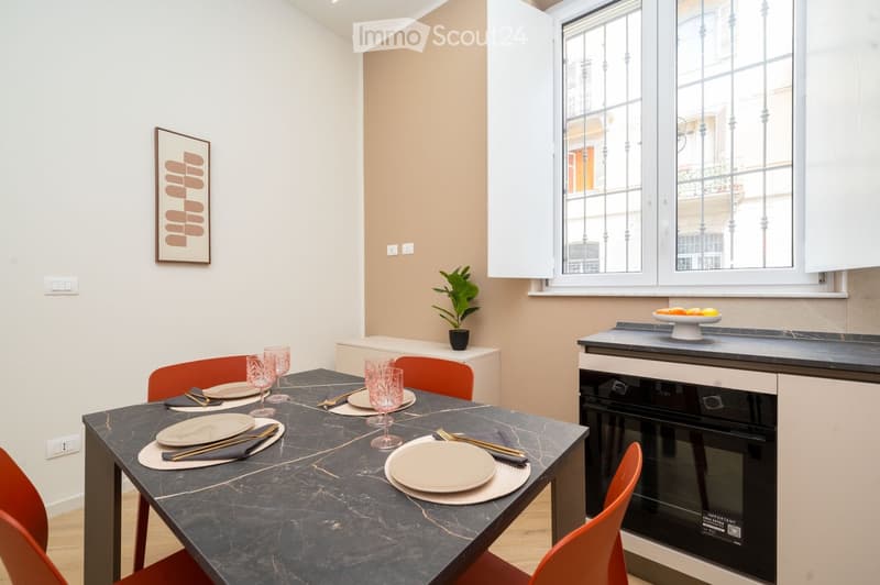 Modern dining area with a dark marble dining table, four orange chairs, wine glasses, plates, and cutlery. A small plant sits on a white cabinet, and a framed picture hangs on the wall. An oven is visible in the background, and a window with decorative ba