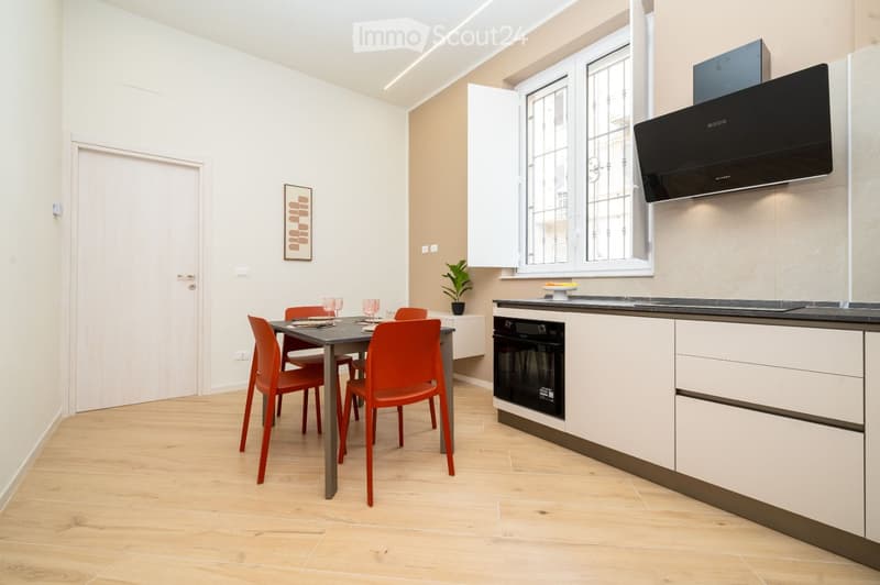 Modern kitchen and dining area with white cabinets, a black oven, a dining table with red chairs, and a potted plant on the windowsill.