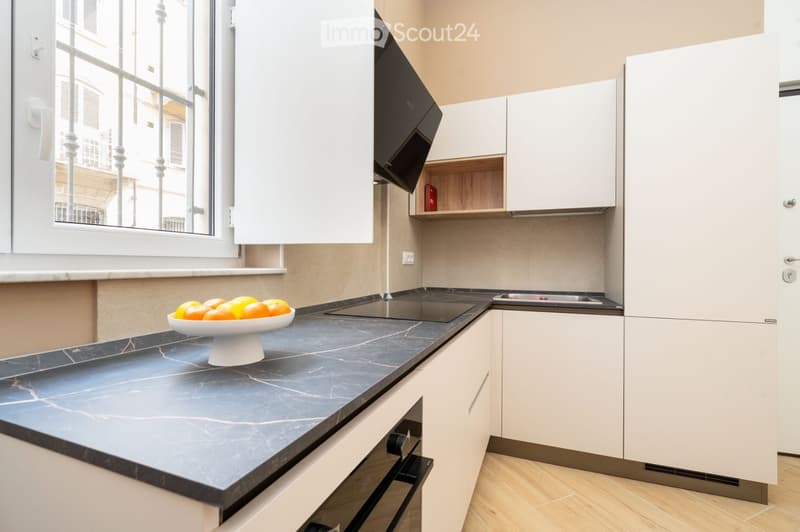 Modern kitchen with white cabinets, dark marble countertop, induction cooktop, and a bowl of oranges on the counter.
