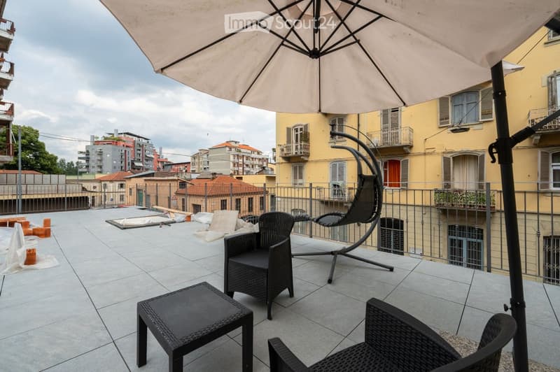 Balcony with tiled floor, black wicker chairs, small table, and large umbrella. Background shows cityscape with buildings and balconies.